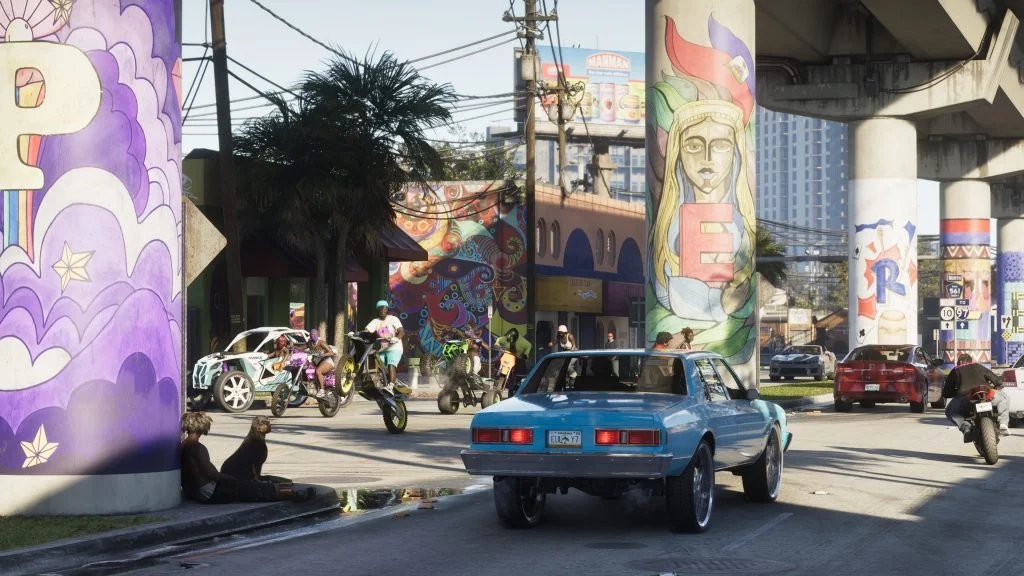 Uma captura de tela vibrante de Grand Theft Auto VI (GTA 6) mostrando uma rua movimentada em Leonida com um grupo de motociclistas e pilotos de quadriciclo realizando acrobacias em frente a murais coloridos. Em primeiro plano, um sedã clássico azul claro dirige-se em direção ao grupo, enquanto um homem e seu cachorro sentam-se na base de um pilar de concreto decorado com grafites. O fundo apresenta um viaduto rodoviário, palmeiras e edifícios modernos da cidade, capturando a atmosfera de um cenário ficcional inspirado em Miami.