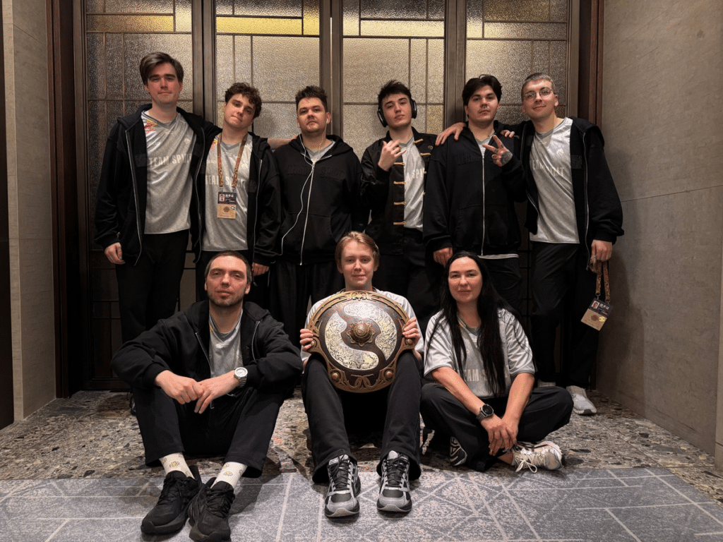 A imagem mostra o Team Spirit posando juntos em frente a uma parede moderna com painéis de madeira, comemorando sua vitória no The International 2026 na China.