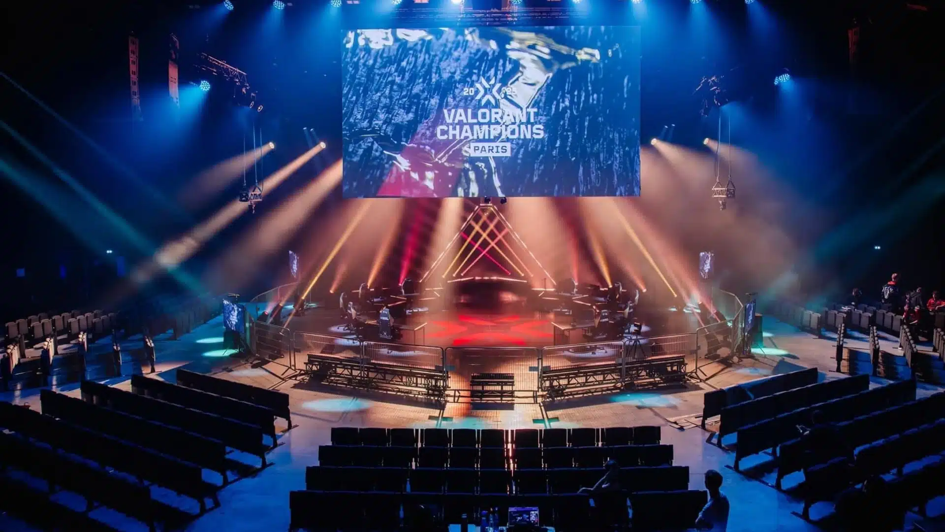 Um palco de esports para VALORANT Champions Paris com telão, iluminação vermelha e azul e assentos vazios.