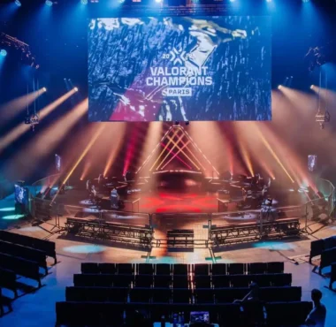 An esports stage for VALORANT Champions Paris with a large screen, red and blue lighting, and empty seating.