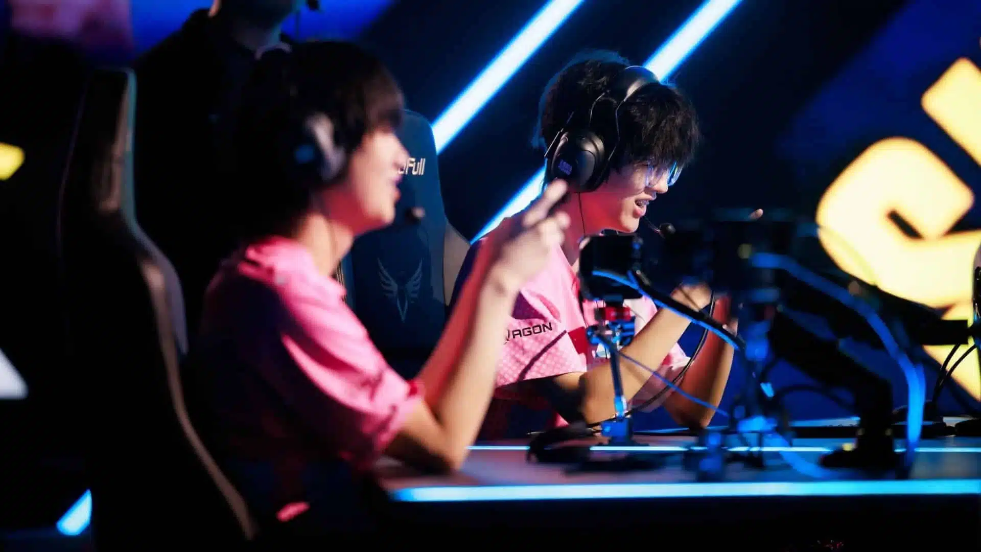 Dois jogadores profissionais de esportes eletrônicos com camisetas rosa e fones de ouvido em uma fase de competição.