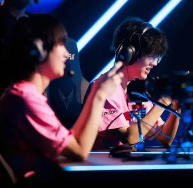 Two professional esports players in pink jerseys wearing headsets at a competition stage.