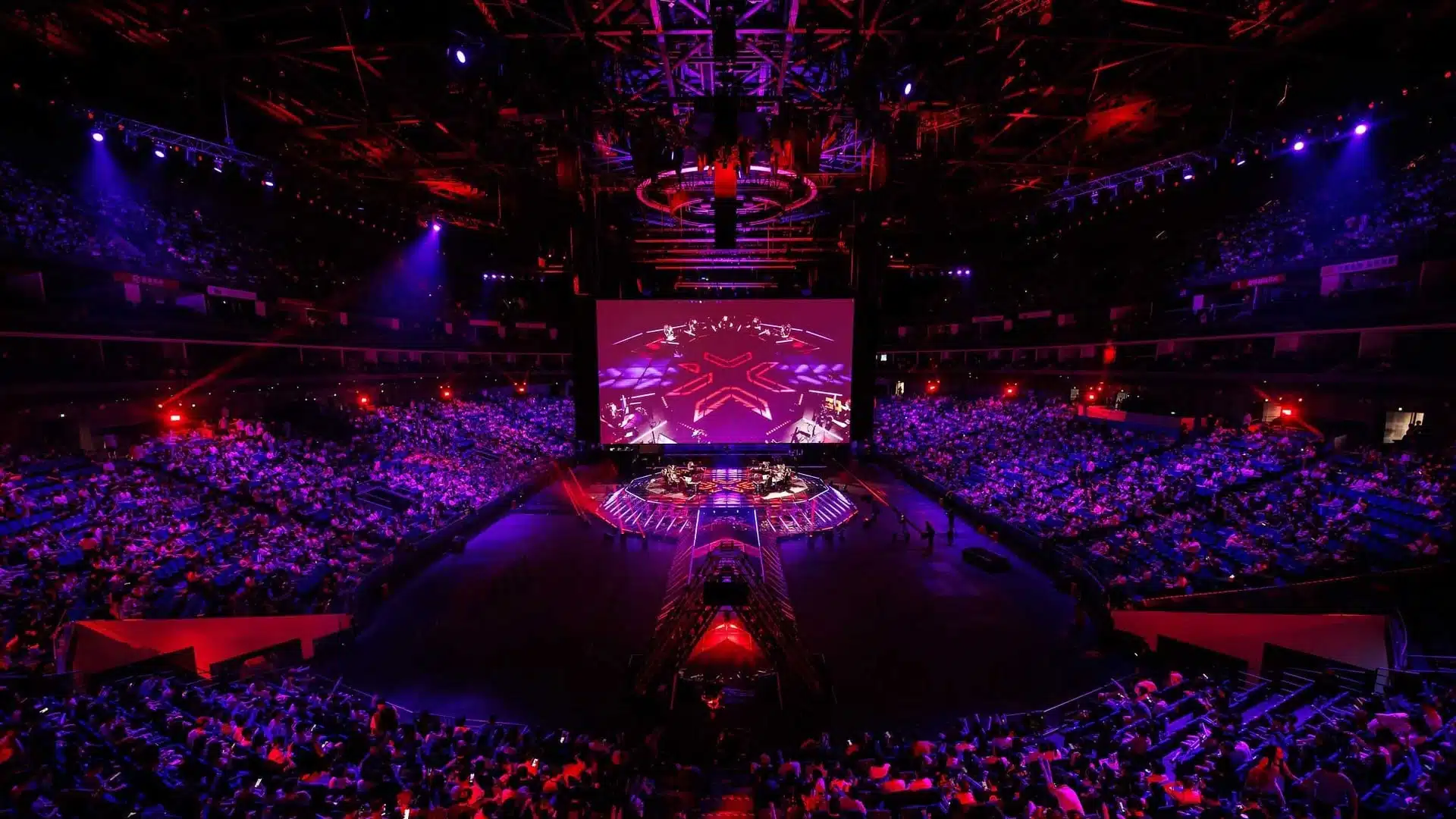Plano geral do palco e público da arena Valorant Champions Shanghai sob iluminação vermelha e roxa.