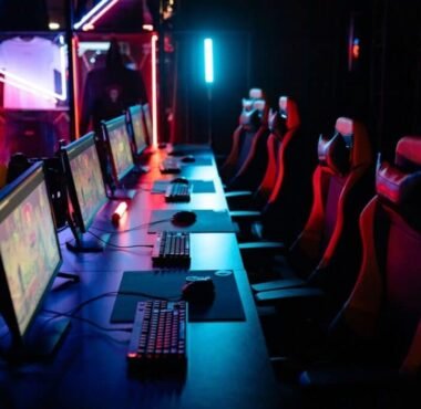 Row of gaming PCs, keyboards and red gaming chairs under neon lighting