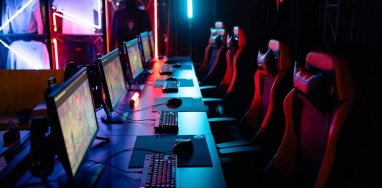 Row of gaming PCs, keyboards and red gaming chairs under neon lighting
