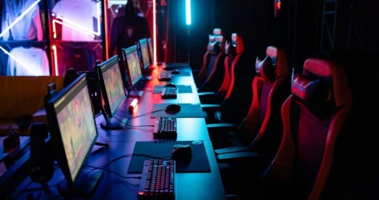 Row of gaming PCs, keyboards and red gaming chairs under neon lighting