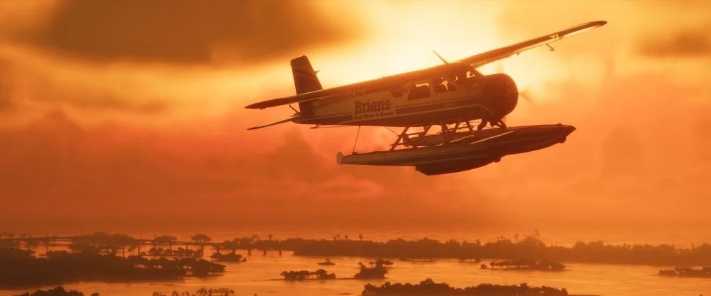 Uma captura de tela panorâmica da hora dourada de GTA VI: um hidroavião voando baixo sobre a água e ilhas tropicais, com a marca "Obras e Marinha do Barco de Brian" visível na lateral da aeronave.  